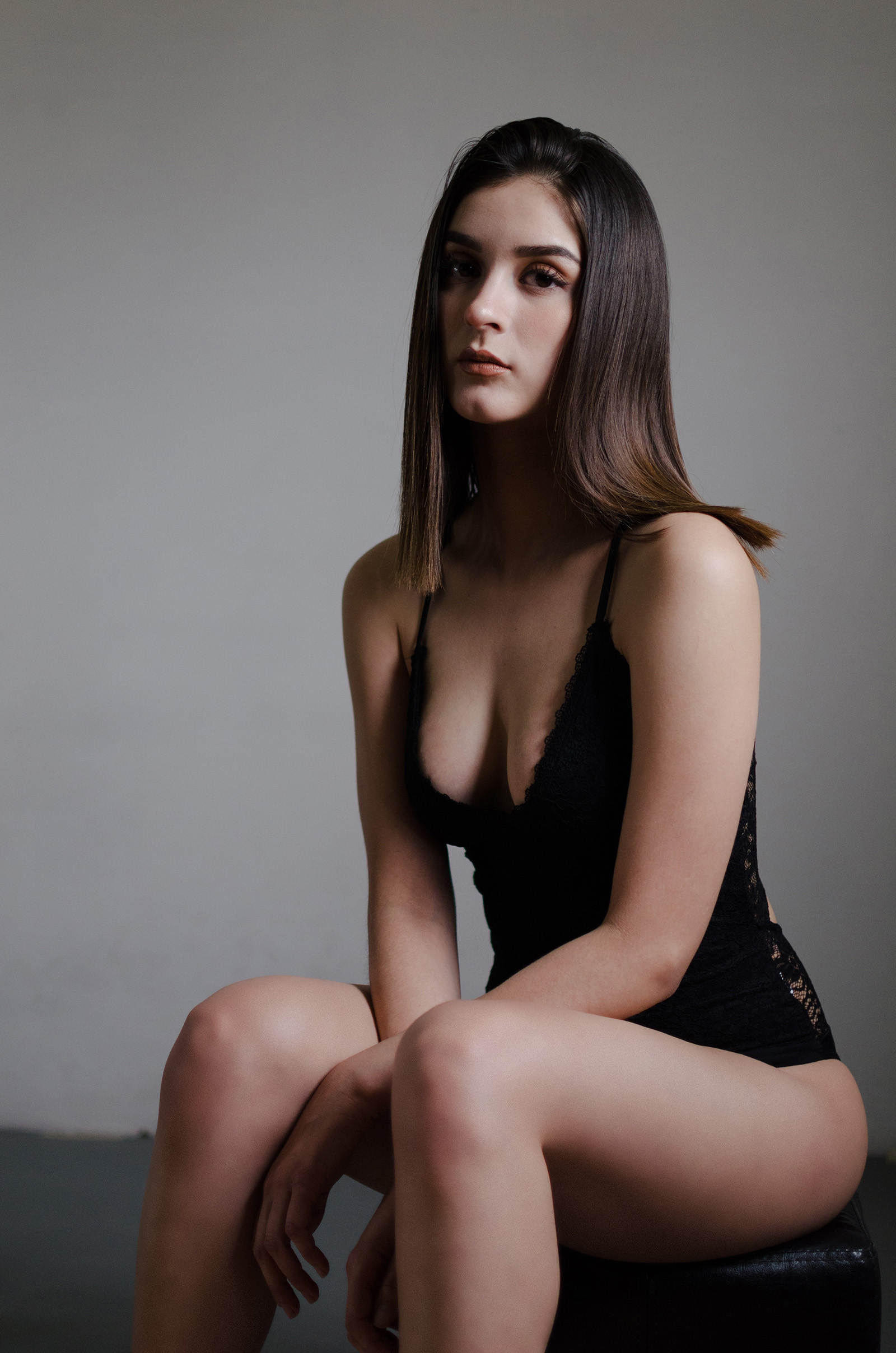 Cassandra Villareal sits against a muted backdrop wearing a black lace bodysuit with her long brown hair cascading down her shoulders.